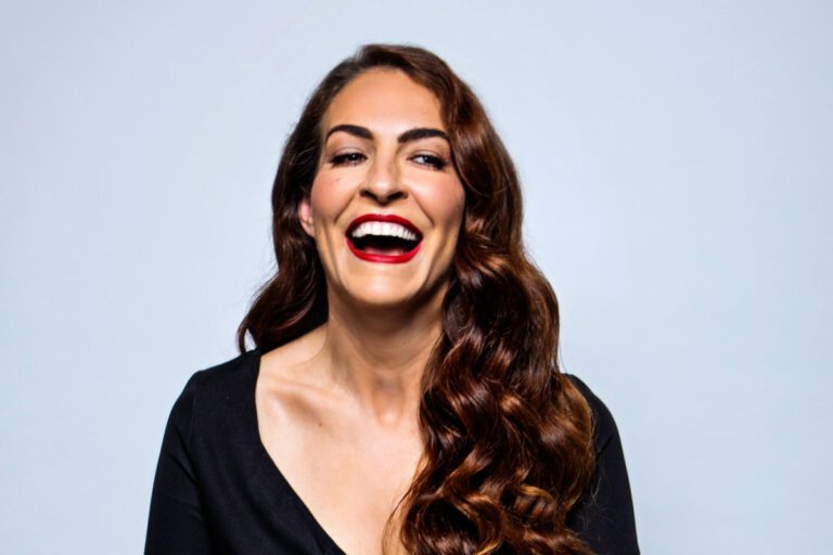 Headshot of Esther Manito laughing against a baby blue background. Esther has long wavy brunette hair. She is wearing a black top.