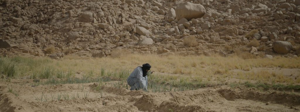 An Arab man is crouched in a sandy coloured desert landscape. He is wearing a black turban
