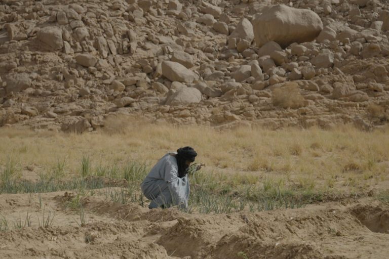 An Arab man is crouched in a sandy coloured desert landscape. He is wearing a black turban