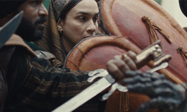Cropped image of warriors protecting a queen using red shields. Swords are drawn. The queen has a tense look on her face in between the shields.