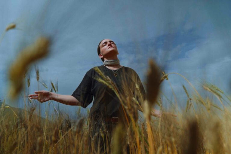 Artist lisa luxx is standing with her arms open in a wheat harvest field. Her eyes are closed and head titled slightly upwards.